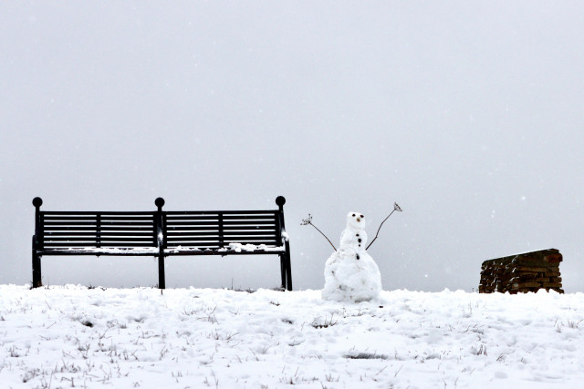 A snowman with long twig arms pointing up stands on the top of a snowy hill between a metal bench and a stone cairn. Grey snow clouds fill the sky behind 