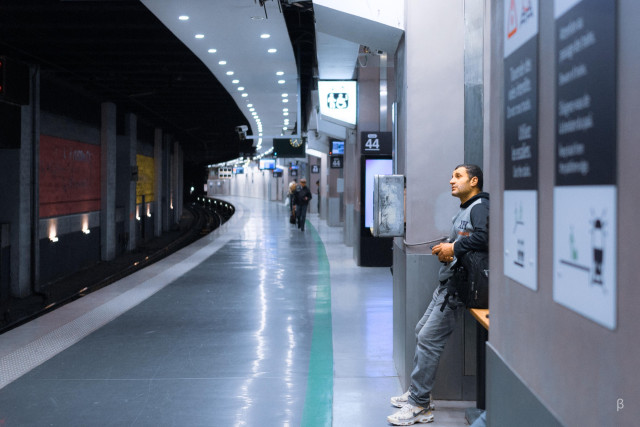 A modern subway platform with a curved design, lit by a line of bright recessed lights that create a vanishing point perspective. The platform has a distinctive green line running along its edge. A person in casual wear - a grey sweatshirt and light-colored pants - leans against a station pillar marked with the number 44. The platform appears largely empty, with just a few distant figures visible in the background. The sleek, modern design features white walls, digital displays, and a polished floor that reflects the overhead lighting.