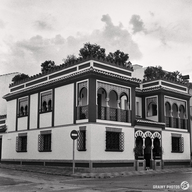 A black-and-white film photo of a Spanish house on a street corner. There is a large balcony on the first floor with ornate arches. Orange trees are growing on the flat roof Terrace.