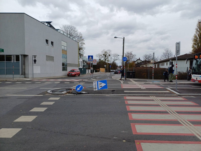 Bild einer Kreuzung am Stadtrand. Die Fotografin steht vor einem Fussgängerübergang mit anschliessender Radfahrerüberfahrt, die sich über zwei in der Mitte geteilte Fahrbahnen erstrecken. Der Fahrbahnteiler ist eine Verkehrsinsel, auf dem eine Stange montiert war, auf der ein Richtungspfeil, ein Hinweis auf den Fussgänger- und Radfahrerüberfahrt und die Zusatztafel "Schulweg" montiert sind. Das Schild liegt waagrecht auf dem Boden, offenbar von einem Auto niedergefahren, Teile des Autos befinden sich daneben. Im Hintergrund ein paar langweilige Stadtrandbauten und ein bissl Grün, das im November natürlich nicht besonders grün ist.