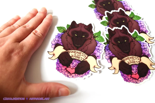 Hand next to a pile of vinyl stickers for scale, which feature a black phase wolf with piercing yellow eyes surrounded by purple-pink hydrangea flowers. A banner draped across the wolf's forelegs reads: "Kindness Is Not Weakness".