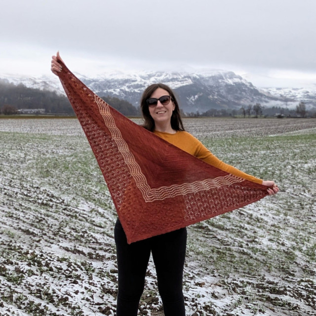 1. A square photo of me holding up my finished hand-knit shawl, outstretched. The shawl is triangular, with MC a rusty brick red colour and CC a dusty pink colour with occasional yellowy and reddish speckles. It has been worked downward and outward from the centre back. The top section is MC, and has a 'dot' texture made of purl bumps interspersed throughout stockinette. The middle section has rows of MC and CC Old Shale lace, that is, a scalloped fan-like lace. The bottom section is MC, and has a pattern of nupps surrounded by simple lace circular shaping. I am smiling at the camera, and wearing a yellow jumper and black jeans. I am standing in a lightly-snowy field, and in the distance are some snow-topped hills.