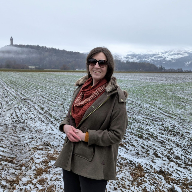 2. A photo of me wearing the shawl wrapped around my neck under a billowy green tweed swing coat with faux-fur collar. I am in the same field and smiling at the camera. The camera angle has moved slightly so as well as the snowy hills behind me there is a tall, pointy (phallic) monument on a small hill.