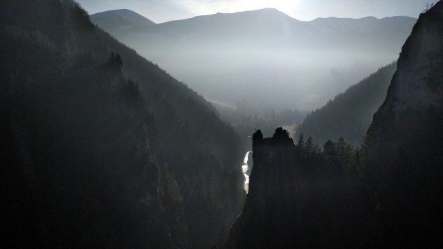 Almost black and white looking photo of a deep narrow valley between rocky, tree-covered hills with other mountains in the background. The foreground parts are in shadow only a thin fog filling the valley is lit by the low sun.
There is a narrow mountain stream flowing through the valley.