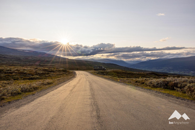 Gravel road in the wide landscapes of Norway. The sun is just setting behind a bank of clouds and makes a sun star. A black van stands at the side of the road.