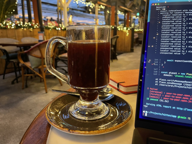 Photo of a bar/restaurant with a sunset visible through the windows at the back. In the foreground is a cup of mulled wine and a partially-visible computer screen showing some failed PlayWright tests.