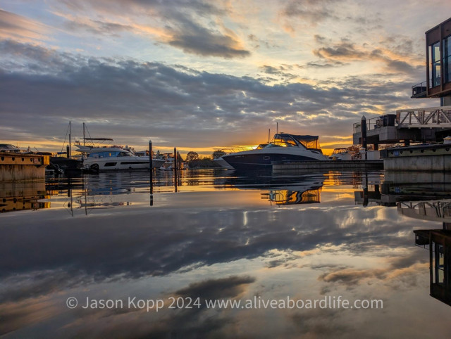 Sunsetting behind 20ish ft cruiser with white deck and red hull. Low gray clouds and some light blue sky behind all reflect in still water at Gangplank Marina