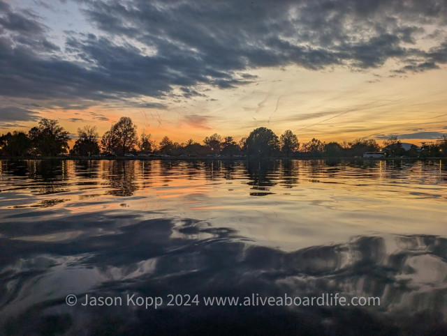 Washington Channel calm water with gentle waves reflects hains pt tree line m, some dark clouds overhead and light pink clouds lower near gold horizon