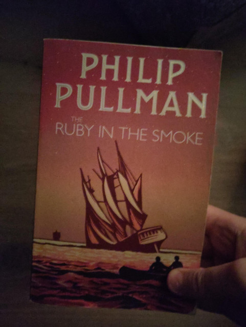 The front cover of The Ruby in the Smoke depicts a sinking ship in sunset colours. In the background, there is another ship so distant you can barely make out its features. In the foreground, there is a small boat with two survivors looking at the main ship sinking.