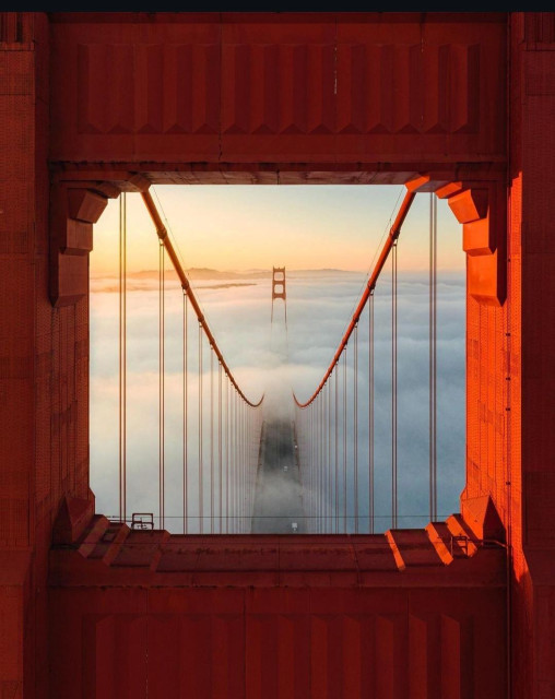 Photography. A color photo of the Golden Gate Bridge in San Francisco. The photo shows a view from above on one of the orange-red bridge girders. One looks down through the bridge girders onto the suspension bridge, which is shrouded in fog. Only the upper part of the bridge and the roadways are visible. The golden sunlight illuminates the scenery from the left and gives a hint of the hills on the horizon.
Info: The Golden Gate Bridge is a 227 m long and 1280 m wide suspension bridge with six lanes and two walkways and cycle paths at the entrance to San Francisco Bay. To be able to take drone shots on site, you need an official flight permit, which is not easy to obtain. Incidentally, the photographer didn't have one.