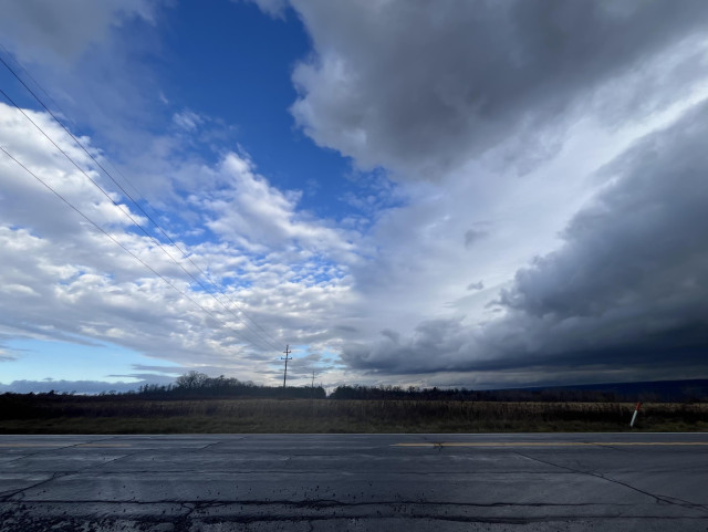 Picture of a road, with a sky behind it, with one half having cumulous clouds and blue skis, and the other half showing dark clouds and rain.