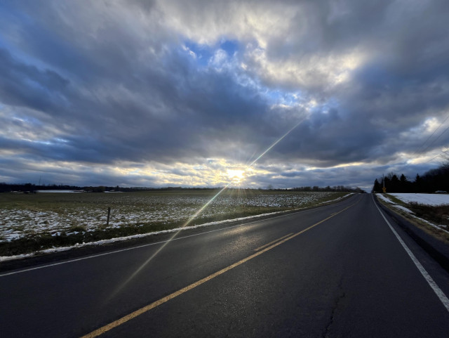 Picture of the sun shining through clouds and reflecting off snowy fields and a moist road.