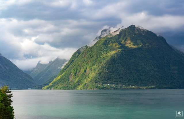 Color photo of sunlit mountains rising steeply from a fjord with clouds on their peaks. 