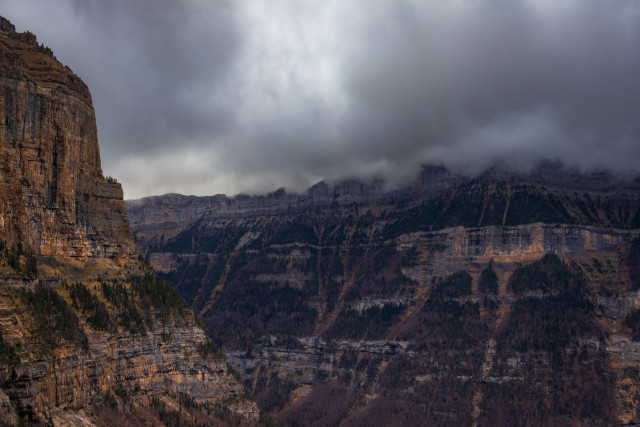 Se ve un paisaje montañoso nublado. Con nubes de posible tormenta. A la izquierda hay un acantilado que marca parte de la entrada al valle con las montañas del fondo, coronadas por nubes.