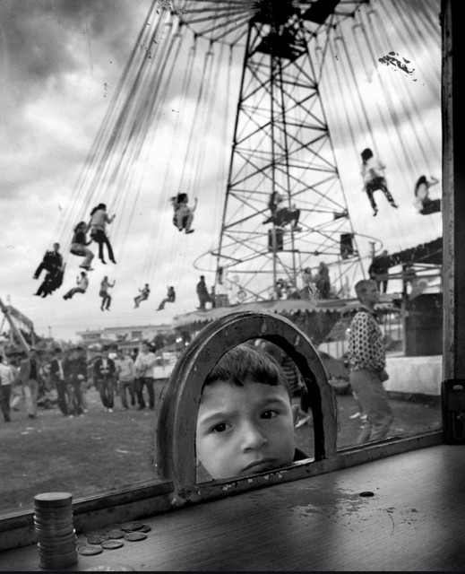 Photography. A black and white photo of a scene at a funfair. It shows a child at the cash desk of a fairground stall. In the foreground is the window of the stall with a small opening through which the head of a small boy can be seen. The photo was taken from the inside. His gaze shows his eager impatience. There are already some coins on the counter. The background is blurred and shows a large chain carousel on which people are swinging in their seats. They are moving fast. Other people are walking across the square. The photo conveys a slightly nostalgic atmosphere and brings back fond childhood memories.