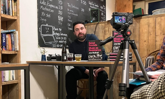 A photo of Joe Mulhall, speaking and playing music at a secret event held in east London on 17 November after a pub became the latest in a string of venues to pull out of events to promote his book.

He is sitting behind a table with a camera aimed at and a blackboard behind him, in what looks like a small coffee/book shop.