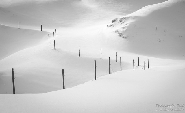 A serene winter scene is captured in this image, showcasing a snow-covered landscape with a fence running through it. The snow is pristine and untouched, creating a peaceful and tranquil atmosphere. The dominant colors in the image are white and grey, highlighting the winter wonderland setting. The image is in black and white, adding to the timeless and classic feel of the scene. The snowy slope and fog in the background add to the mystery and beauty of the setting. This picturesque setting is perfect for nature lovers and those who appreciate the beauty of winter landscapes.