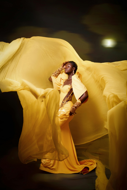 A Black fem in an ornate golden-yellow gown and long gloves, with yellow fabric blowing all around her, against a dark background. 