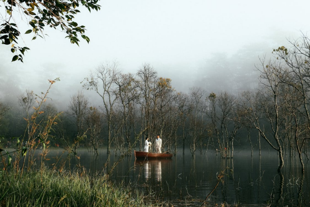 Two people in white standing in a little boat on still waters, with misty trees in the background and a bank of long grass in the foreground