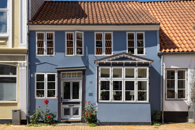 A blue house nestled in between two other buildings, with a terracotta tiled roof, white-panelled wood windows, and red flower bushes growing outside. 