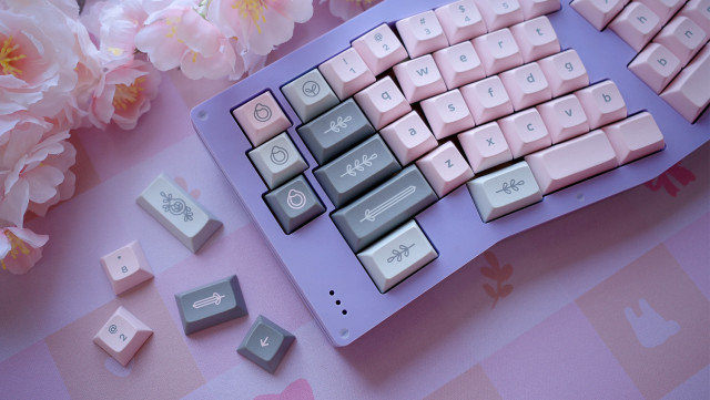 Angled view of a lavender colored keyboard with light blush pink and green keycaps on a picnic themed deskmat that matches.