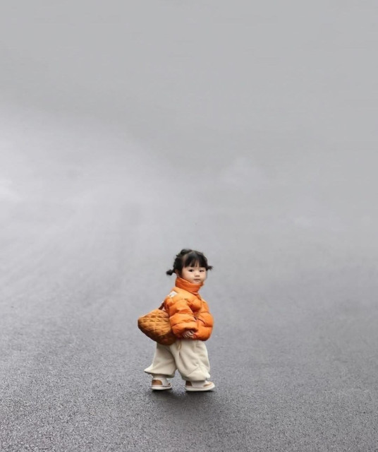 Photography. A color photo of a little Asian girl in winter clothes. She has little pigtails and is wearing a thick orange down jacket, a bag and beige bloomers. Three quarters of the picture consists of a gray street, which makes the little fashion-conscious girl look absolutely tiny and cute. 
Info: The photo is a fashion photo from the Korean children's fashion company Ainini.