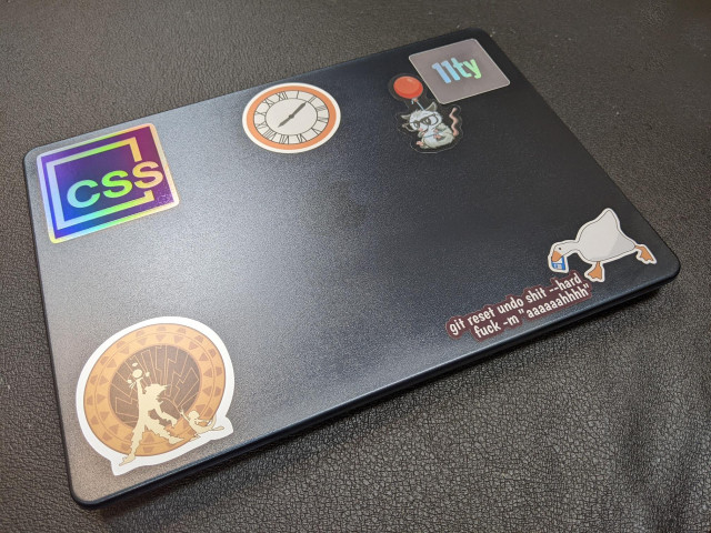 A laptop with stickers on it: a holographic CSS one, a clock from Back to the Future, a drawn opossum hanging on a balloon next to an Eleventy logo, a Jak & Daxter silhouette, a fake panicked git command, and a goose running with a floppy disk in its beak.