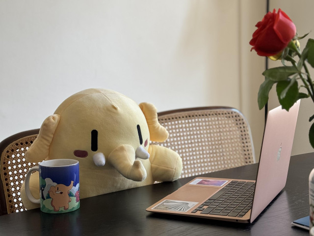 A Mastodon Plushie sitting at a table in front of a laptop with a Mastodon mug