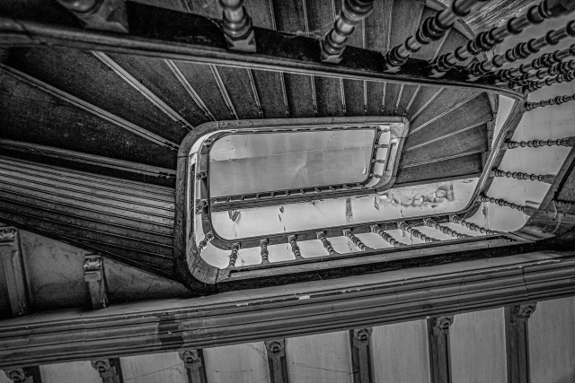 Das Schwarz-Weiß-Foto zeigt eine Wendeltreppe aus dunklem Holz, die sich nach oben schraubt. Die Treppe ist detailreich gearbeitet, mit geschwungenen Geländern und kunstvollen Verzierungen.  Durch eine Öffnung in der Treppe ist ein weiterer Abschnitt der Treppe sichtbar, der sich in die Höhe windet. Der Gesamteindruck ist der eines alten, möglicherweise historischen Gebäudes mit einer beeindruckenden Treppenkonstruktion