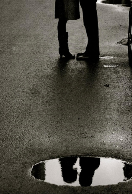 Photography. A black and white photo of a couple next to a puddle of rain with an interesting composition. The very dark photo shows an asphalt road with a puddle of rain in the foreground. The upper part shows the feet and legs of a couple facing each other. The woman's boots and skirt, the man's trousers and boots. In the puddle below, the shadow outlines of the two heads can be seen as they are very close to each other. A beautiful way to depict the theme of love with black and white photography