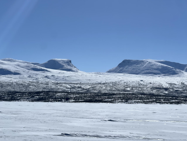 An almost all white landscape, flat frozen lake with snow on top in the foreground followed by some sparse trees following its shoreline. There’s a flat mountain range in the background with a very prominent U valley at the center, almost like a gigantic halfpipe. Bright blue sky. 