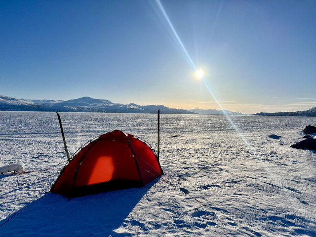 Red dome style tent in the foreground. Two skis pitched left and right to serve as stakes. View is across the snow covered, frozen lake towards a bright sun about to set, faint mountains at the very distant horizon. Blue sky, not a single cloud. 