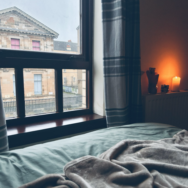 A square photo in colour shows a scene looking out the window, in Glasgow. In the foreground is the edge of a bed with a soft blanket on top. In the mid-ground is the windowsill in dark wood with a shiny finish that is a bit reflective from the light coming in the window, giving the wood a glossy sheen. Above the windowsill, still to the left of the frame, is the window, out of which can be seen an old yet well-maintained sandstone building with a steepled rooftop that points towards the sky. The sky is a pale grey-blue of an approaching storm. Inside the room, to the right of the frame, is a white pillar-candle that sits in its candleholder on top of a white radiator. The white wall behind the radiator, & the candle itself, appear to glow a deep yellow-orange from the contrast of the blueish light from outside. The photo has a calm & peaceful quality.