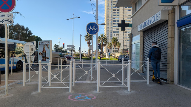 Photo de jour, un large trottoir est pratiquement entièrement bloqué par des barrières. Au sol, un logo fauteuil roulant bleu et un logo de stationnement interdit rouge. Sur la gauche, un petit passage, un homme en trotinette électrique passe. 