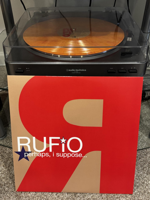 A vinyl record player is playing an orange and red vinyl record. In front of the player is a record cover for the album "perhaps, i suppose..." by Rufio, featuring a prominent red 'R' design.