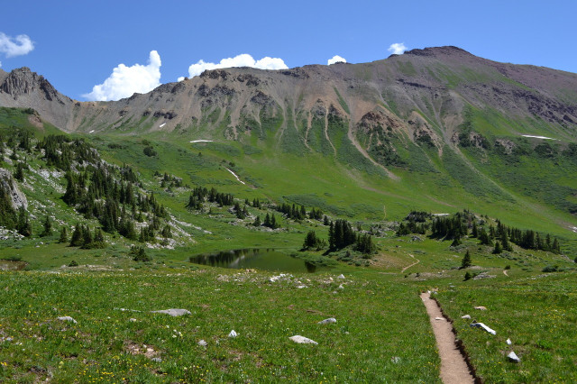 Very distinct dirt path through green covered flat area with a pond in the middle. Scattered trees (conifers) of a perfect tall triangle shape cluster on a gentle slope rising left of the pond. In the distance, a much steeper slope rises, leaving the green behind. It is studded with exposed rock outcrops among loose rock slopes.