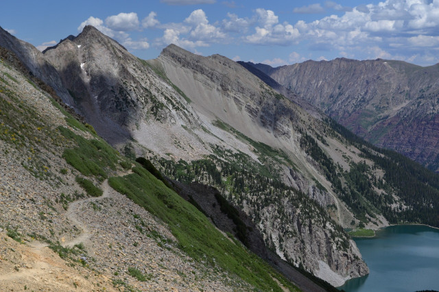 Overlooking Snowmass Lake

A large lake far below the viewer on the right has steep mountains rising to the left and beyond. A trail winds a flat way across this steep slope. A few puffs of cloud occupy half the otherwise blue sky.