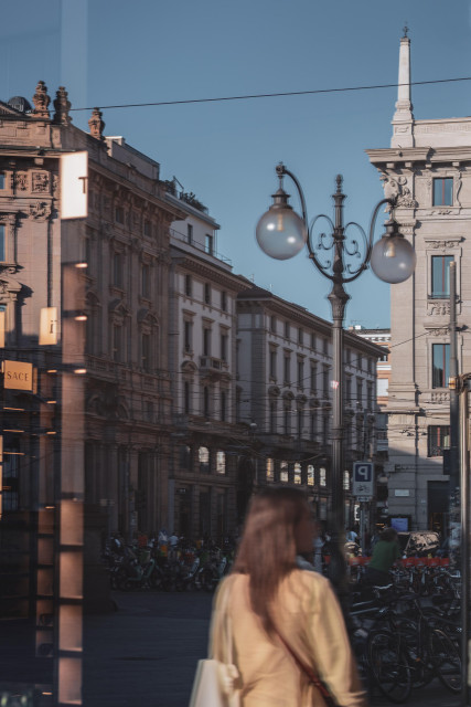 Se observa una vista de calles de Milán, con el sol del atardecer de junio y una chica de espaldas paseando en primer plano. Todo ello captado mediante el reflejo de un escaparate.