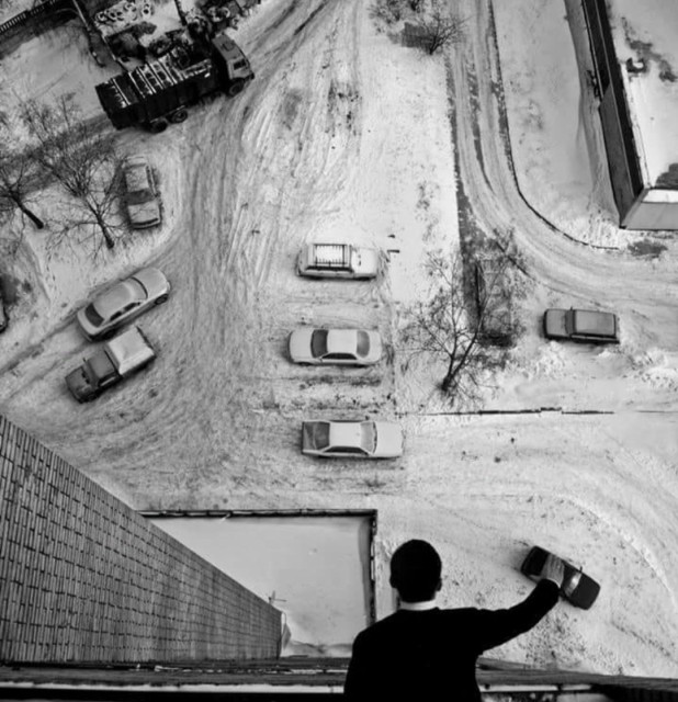 Photography. A black and white photo with a view from above from a balcony onto a snow-covered street with a very funny perspective. A man in black clothing can be seen from above, leaning over the railing of a balcony. Below on the street, moving and parked vehicles can be seen in the snow. The man on the balcony has raised his arm and is covering a dark car on the street below with his hand. The perspective from above makes it look as if he is playing with a small toy car.