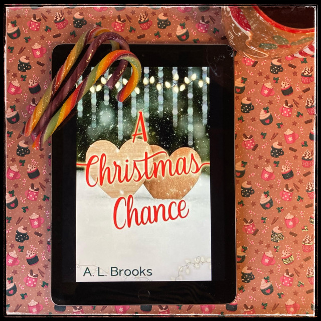 On a festive Christmas-y background, an iPad with the cover of A Christmas Chance by A.L. Brooks. Three multicolored candy canes are set on one corner of the iPad, and there's a gingerbread man mug on the other side.