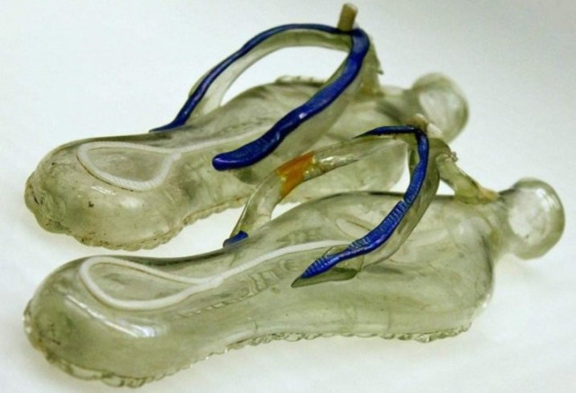 The picture shows flask in the shape of a pair of thonged sandals made of colorless glass with a greenish tinge. The straps are decorated with blue glass.