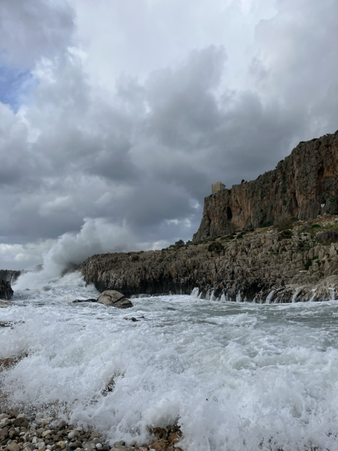 The photo features the Torre di Avvistamento perched on a rocky cliff. Below, powerful waves break against the coastline, enhanced by the dramatic sky filled with heavy clouds.