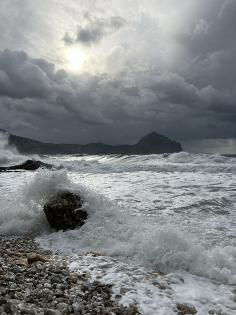 The photo shows Monte Cofano rising prominently in the background. In the foreground, large waves crash against the rocky shoreline, while the sun casts a muted glow through dense clouds.