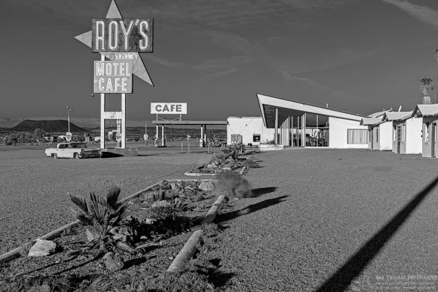 Black and white landscape photo of an abandoned travel center from the 1950s. A large sign on the left with a arrow pointing at an angle toward the ground declares: "Roy's Vacancy Motel Cafe." Further back is a covered gas pump area with a second sign above it that says, "Cafe." Running up the center of the photo is a landscaped island dividing the gravel parking lot from a couple of individual motel cabins. There are some foot to two foot tall palm trees in the islands. A large glass front lobby building is seen at the fall end of the island. The peak of the roof points up towards the large Roy's sign on the left. The sky is dark in contrast to the bright sunlit white buildings. 