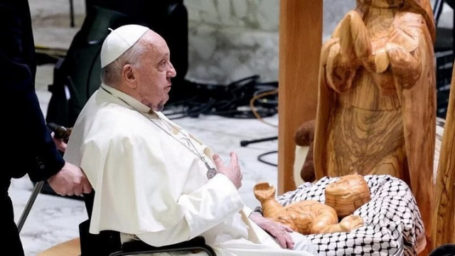 Pope Francis in a wheelchair, in front of a creche made of polished golden wood.