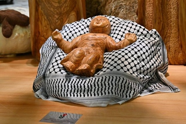 Figure of the Baby Jesus made of polished  golden wood, his arms outstretched to the sides, lying on a black and white keffiyeh 