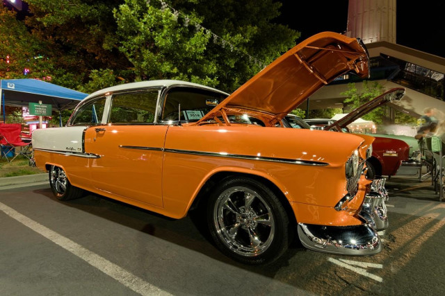 Color landscape photo of 1950s era Chevy Bel Aire car. It is bright orange and white on the rear fenders and top. It has shinny chrome mag wheels. It is on display with the hood open. The photo is from the right front side of the car near ground level. It is night time and bright lights are reflected in the fresh paint. Green trees are in the background.