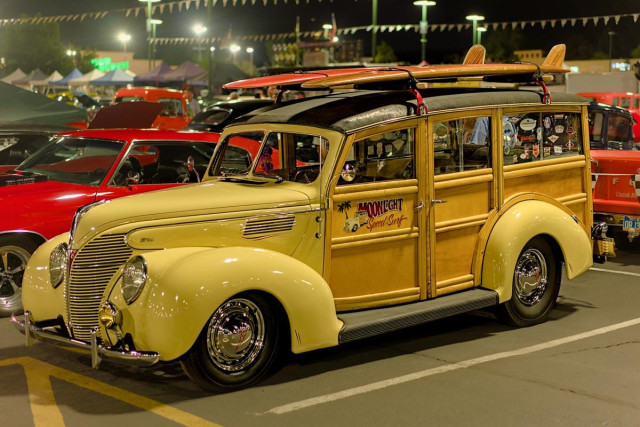 Color landscape photo of 1930s era Ford "Woody" station wagon. It is a night time photo. The vehicle is a pale yellow with wood paneling and a dark leather top. The vehicle has two 1960s style long surfboards strapped to the top. It sits in a brightly lit parking lot with other restored classic cars. 