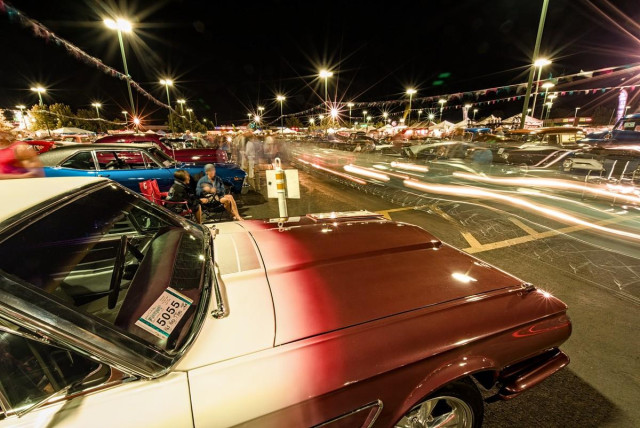 A color long exposure landscape photo of a bright lit parking lot at night. Many classic cars are parked in the lot and the blurred images of people are sitting and standing around looking at cars. There are long white light streams of headlights moving past the camera as people cruise the parking lot. 