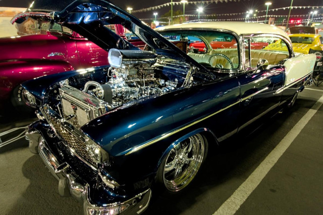 A color landscape photo of a navy blue and white 1950s era Chevy Bel Aire. The front part is blue and the top of the rear fender and top are white. The chrome is very shinny. The hood is open exposing a super charger with a oval air scoop. It is night time and the parking loot is brightly lit. 